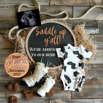 Baby announcement sign with cow print onesie and baby booties on a rustic wooden background